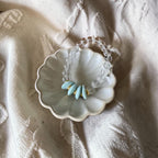 A beaded bracelet with clear faceted beads and five light blue teardrop-shaped beads separated by small gold spacers, displayed on a small white ceramic dish over a textured cream fabric background.