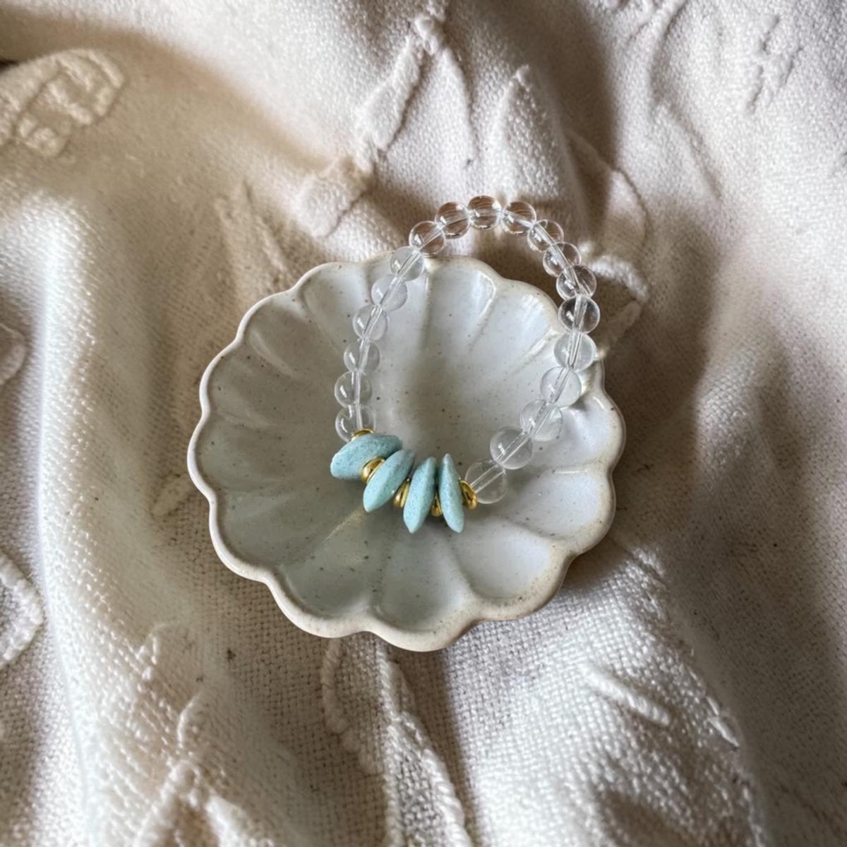 A beaded bracelet with clear faceted beads and five light blue teardrop-shaped beads separated by small gold spacers, displayed on a small white ceramic dish over a textured cream fabric background.