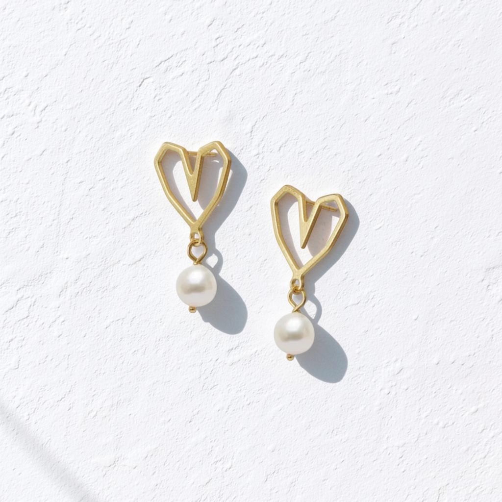A pair of gold heart-shaped dangle earrings with white pearl drops, displayed on a textured white surface with soft shadows.
