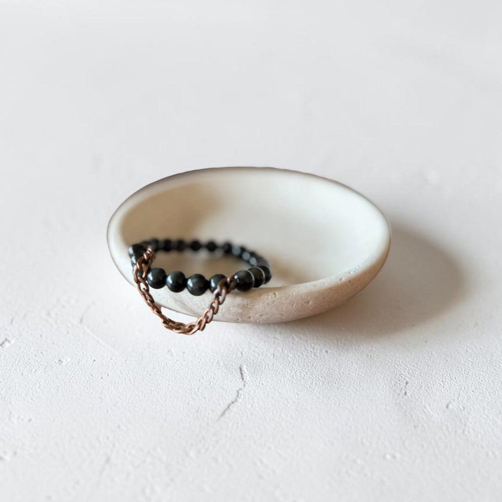 A beaded bracelet with dark round beads and a copper chain link detail, displayed in a small beige ceramic dish on a textured white surface.
