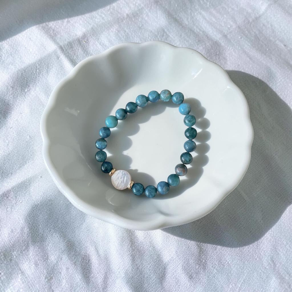 A beaded stretch bracelet with blue-green round beads and a white oval stone charm, displayed on a white scalloped dish over a white fabric background.
