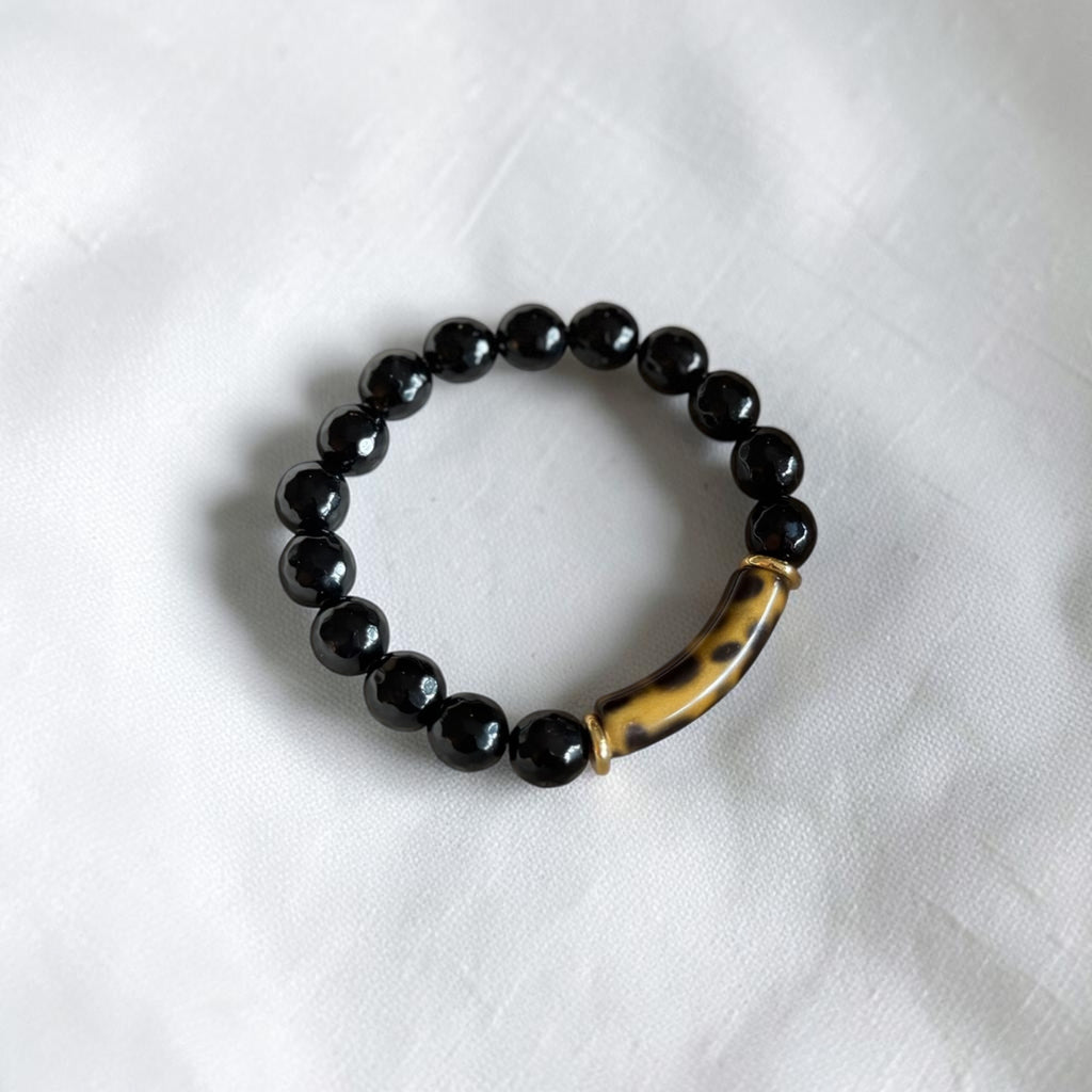 A beaded stretch bracelet featuring glossy black round beads and a curved, polished brown and black tiger-eye style accent bead, displayed on a textured white fabric background.