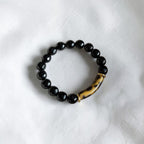 A beaded stretch bracelet featuring glossy black round beads and a curved, polished brown and black tiger-eye style accent bead, displayed on a textured white fabric background.