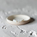 A beaded bracelet with white pearl-like beads and a central clear faceted crystal, displayed inside a shallow cream-colored ceramic dish on a textured light gray surface.