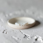 A beaded bracelet with white pearl-like beads and a central clear faceted crystal, displayed inside a shallow cream-colored ceramic dish on a textured light gray surface.