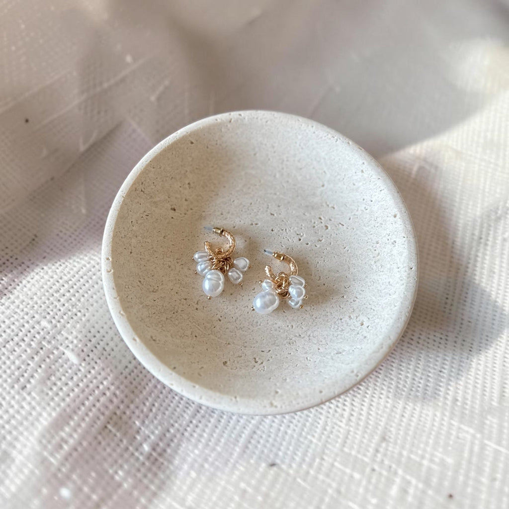 A pair of small gold hoop earrings with white pearl flower clusters, displayed on a small round ceramic dish over a textured white fabric background.