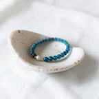 A beaded stretch bracelet with blue and teal beads, a small white bead, and a gold spacer, displayed in a small speckled ceramic dish on a white fabric background.