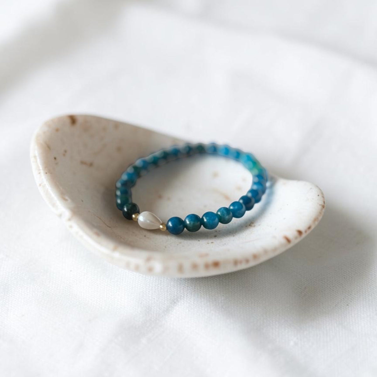 A beaded stretch bracelet with blue and teal beads, a small white bead, and a gold spacer, displayed in a small speckled ceramic dish on a white fabric background.
