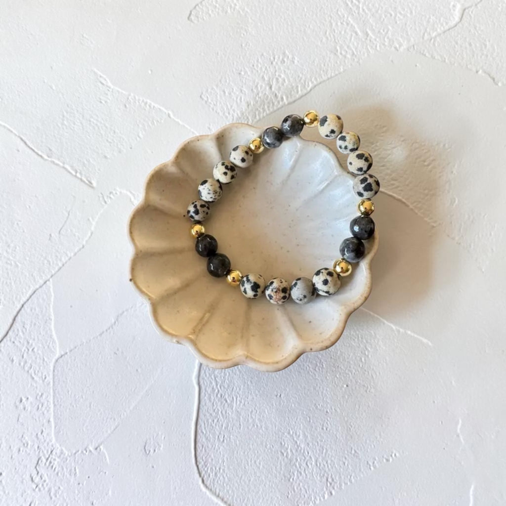A beaded stretch bracelet with black, white, and gray spotted beads and small gold accent beads, displayed on a small scalloped ceramic dish against a textured off-white background.
