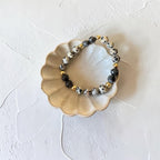 A beaded stretch bracelet with black, white, and gray spotted beads and small gold accent beads, displayed on a small scalloped ceramic dish against a textured off-white background.