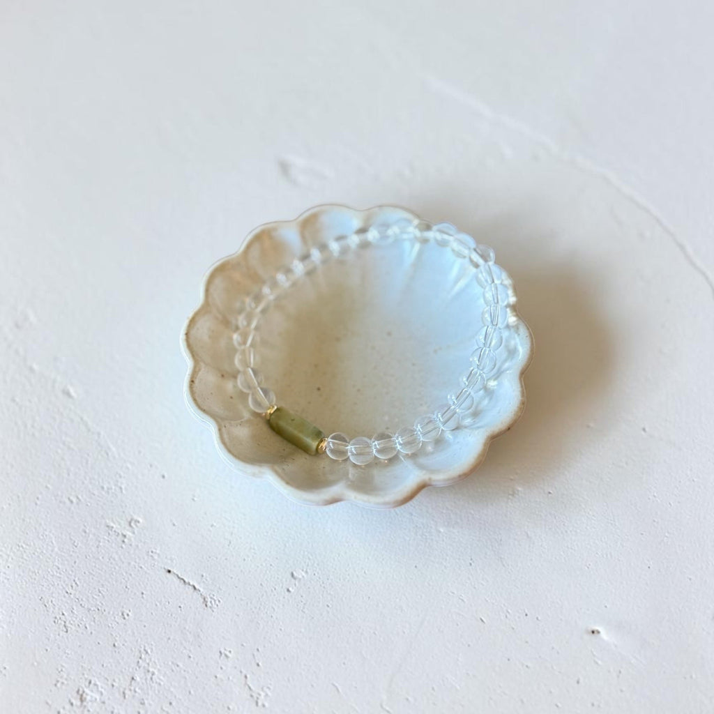 A delicate beaded bracelet with clear faceted beads and a small gold spacer, displayed inside a small scalloped white ceramic dish on a textured off-white surface.