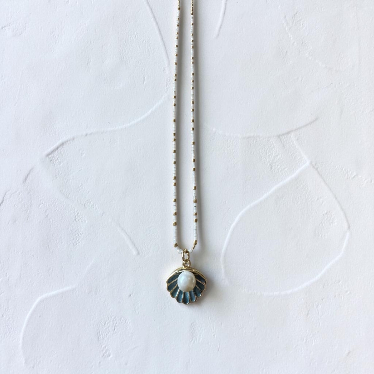 A delicate necklace with a scallop-shaped pendant featuring a white pearl center and blue-green enamel rays, suspended on a fine gold-toned chain against a textured white background.