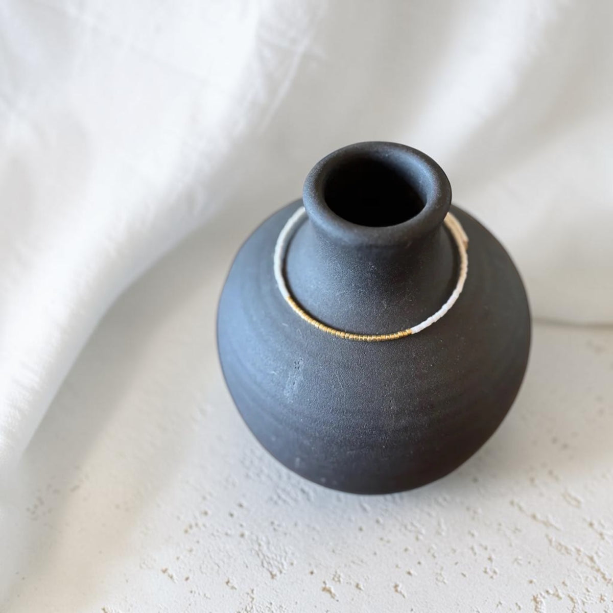 A small matte black ceramic vase with a flared neck and a thin white and bracelet around the upper body, photographed on a light textured surface with draped white fabric in the background.