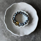 A beaded bracelet with dark blue-gray round beads and a single metallic gold accent bead, displayed on a small white ceramic dish over a larger gray plate.