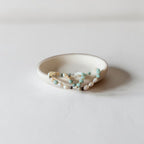 A delicate beaded strand bracelet with white pearls, light blue beads, and tiny gold and black accents, resting inside a small, matte off-white ceramic dish on a light gray surface.