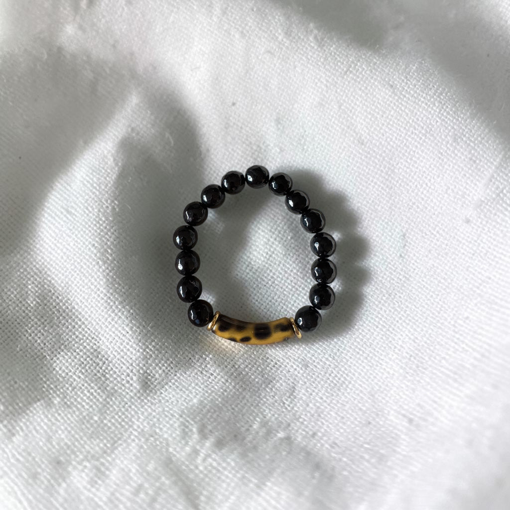 A beaded stretch bracelet featuring glossy black round beads and a central gold-toned cylindrical bead with a black leopard print, resting on a textured white fabric background.
