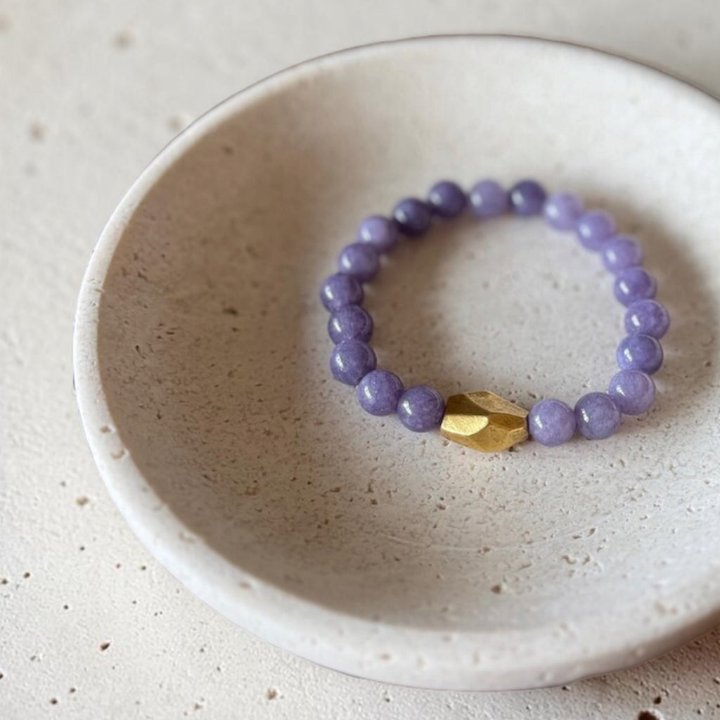 A beaded stretch bracelet with lavender purple round beads and a single faceted gold accent bead, displayed inside a small speckled ceramic dish on a light stone surface.