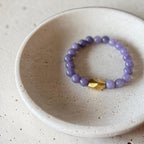 A beaded stretch bracelet with lavender purple round beads and a single faceted gold accent bead, displayed inside a small speckled ceramic dish on a light stone surface.