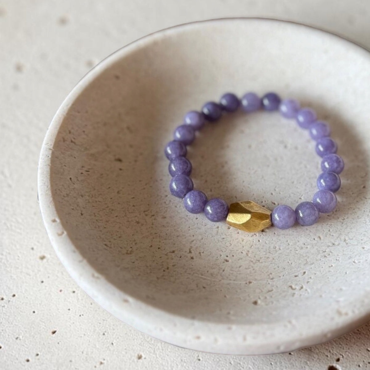 A beaded stretch bracelet with lavender purple round beads and a single faceted gold accent bead, displayed inside a small speckled ceramic dish on a light stone surface.