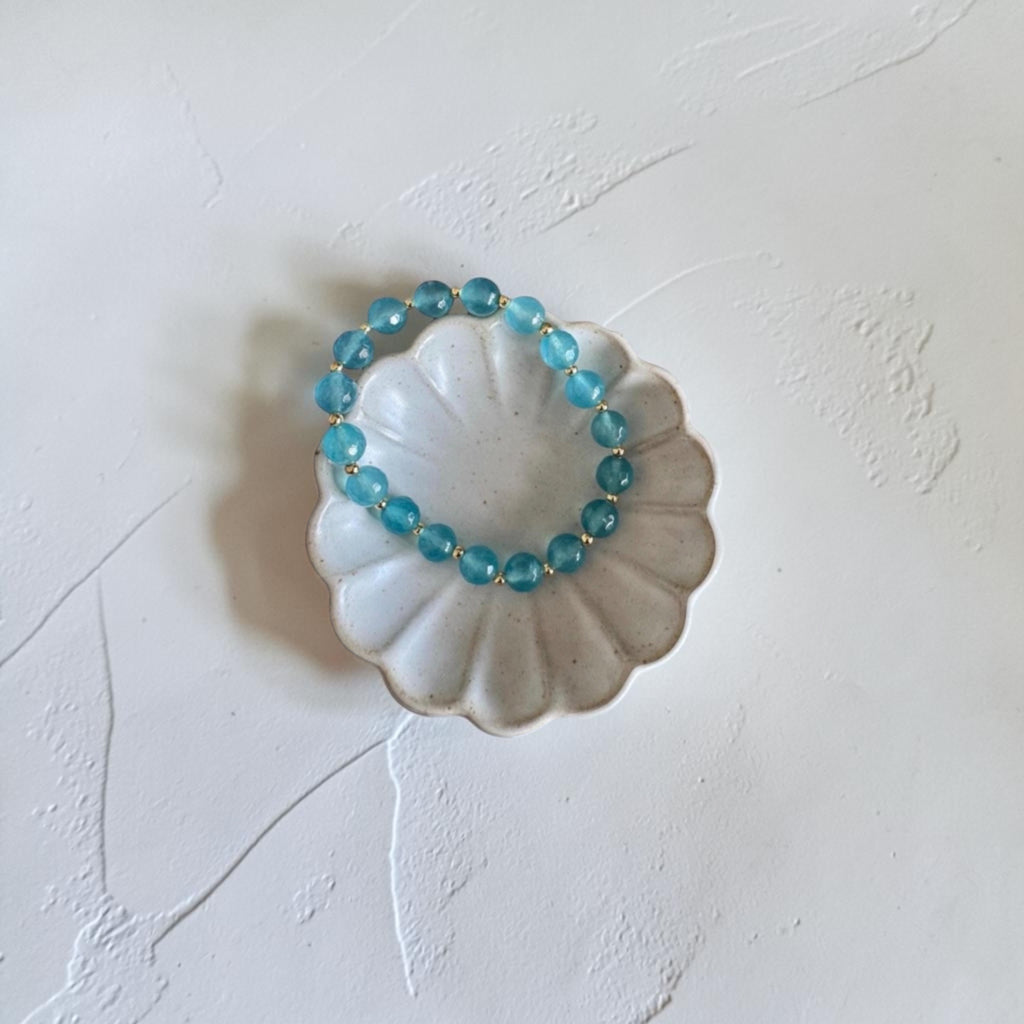 A beaded stretch bracelet made of translucent light blue beads, displayed on a small white scalloped dish against a textured off-white background.