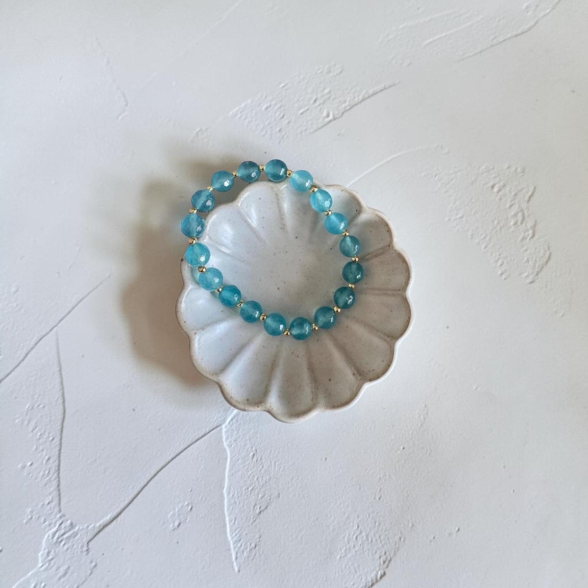A beaded stretch bracelet made of translucent light blue beads, displayed on a small white scalloped dish against a textured off-white background.