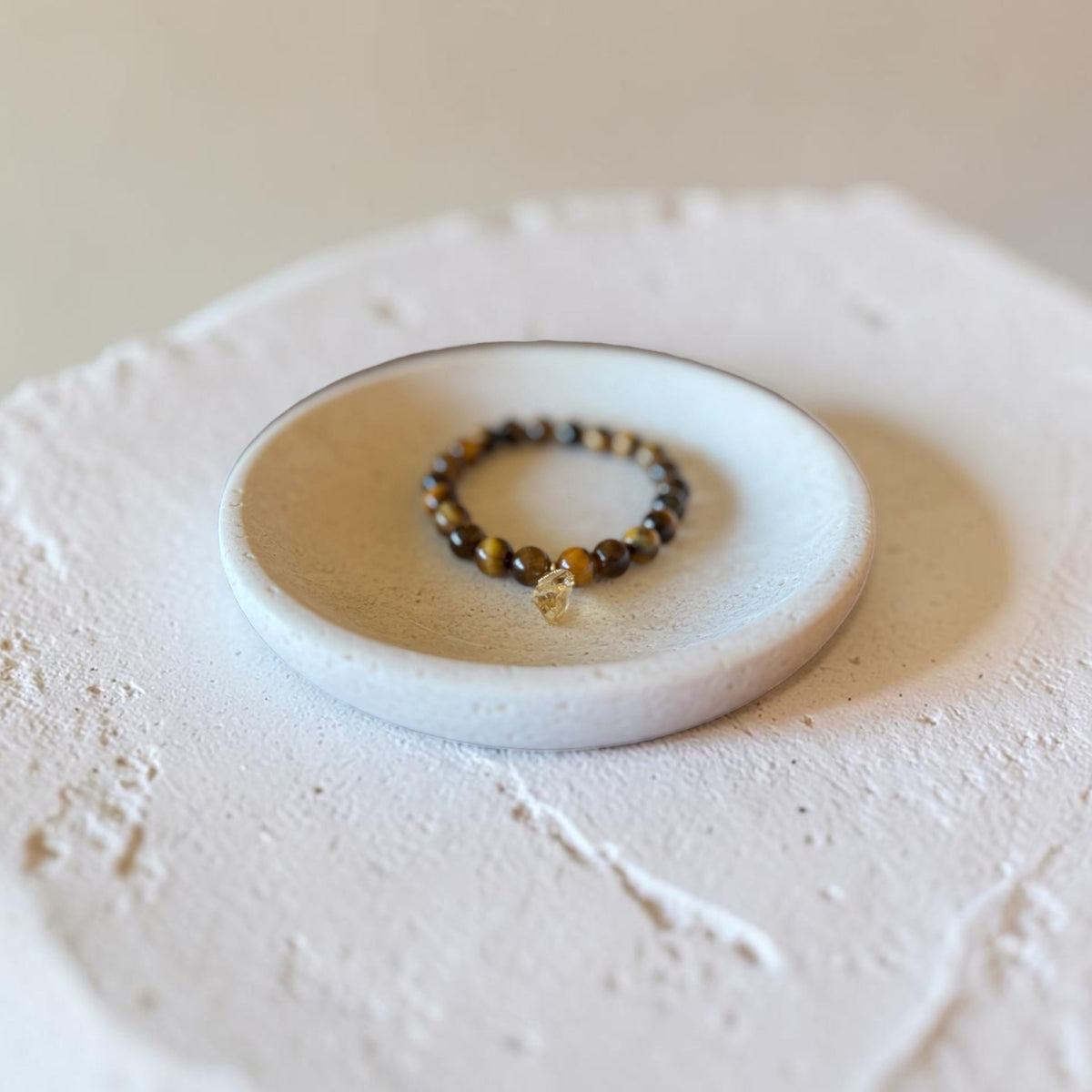 A beaded bracelet with dark brown and black beads and a small clear crystal charm, displayed on a small white ceramic dish over a textured white surface.