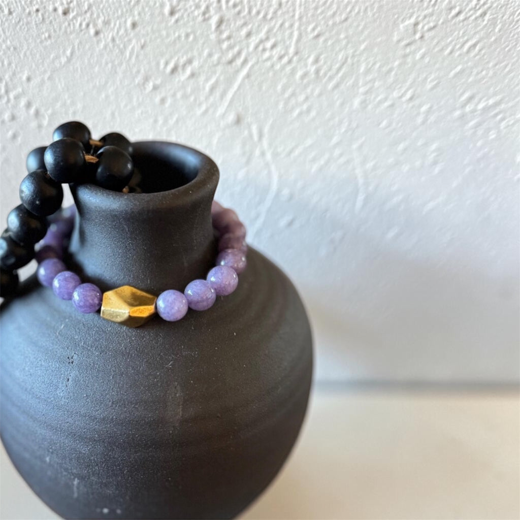 A beaded bracelet with light purple round beads and a faceted gold accent bead, draped over a matte black ceramic vase against a textured off-white wall.