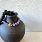 A beaded bracelet with light purple round beads and a faceted gold accent bead, draped over a matte black ceramic vase against a textured off-white wall.