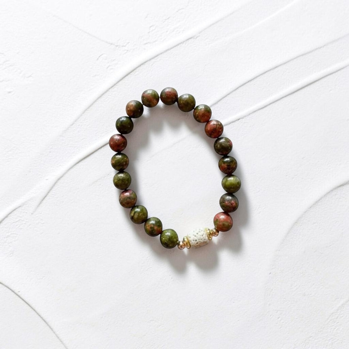 A beaded stretch bracelet made of round green and reddish-brown beads with a small white shell accent, displayed on a textured white background.