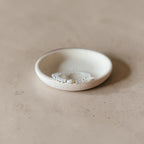 A delicate clear quartz bracelet with a small teardrop pearl its inside a small, shallow, textured off-white ceramic dish on a warm beige surface.