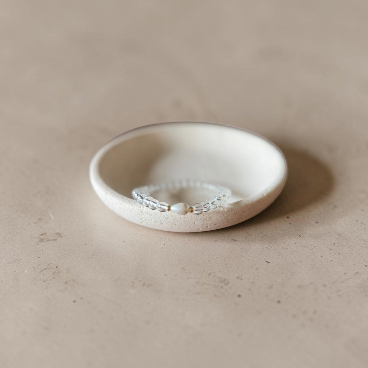 A delicate clear quartz bracelet with a small teardrop pearl its inside a small, shallow, textured off-white ceramic dish on a warm beige surface.