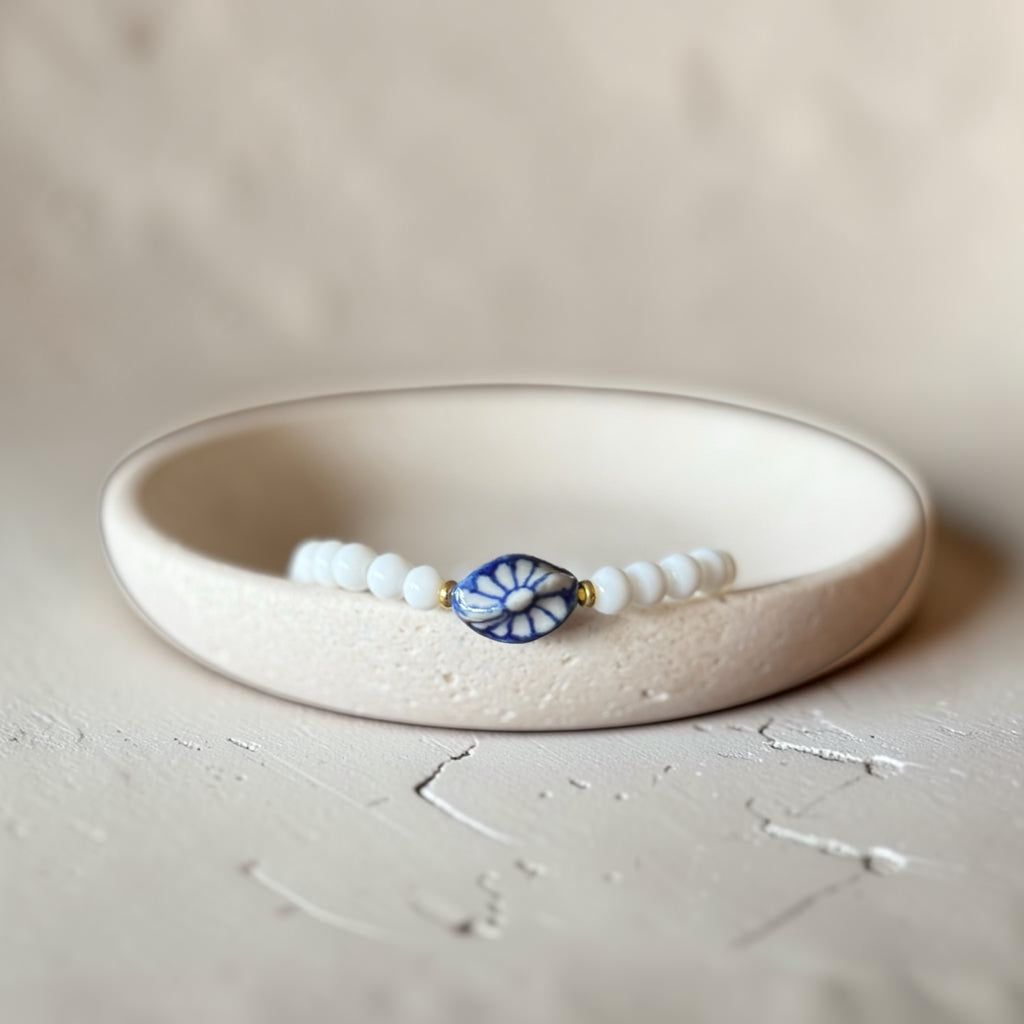 A beaded bracelet with white round beads and a central blue and white floral medallion, displayed inside a cream-colored ceramic bowl on a textured beige surface.