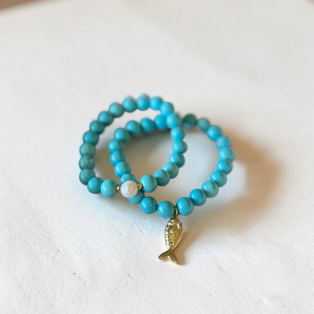 Two turquoise beaded stretch bracelets with a small gold fish charm and a white pearl accent, displayed on a textured white surface.
