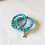 Two turquoise beaded stretch bracelets with a small gold fish charm and a white pearl accent, displayed on a textured white surface.