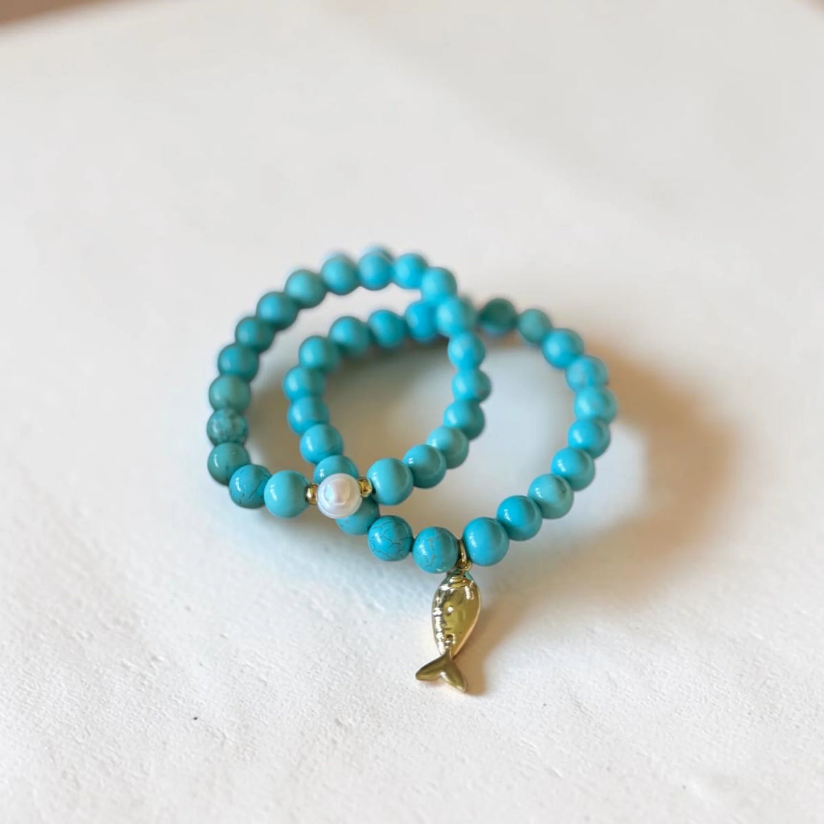 Two turquoise beaded stretch bracelets with a small gold fish charm and a white pearl accent, displayed on a textured white surface.
