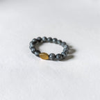 A beaded stretch bracelet featuring dark gray stone beads and a single textured gold accent bead, displayed on a clean white textured surface.