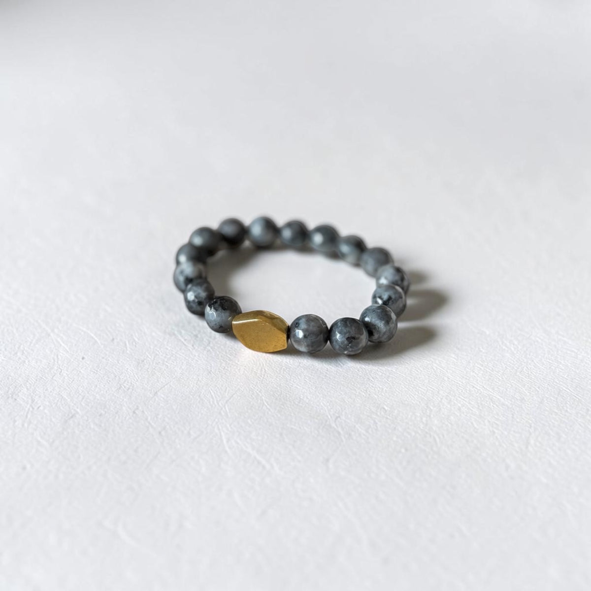 A beaded stretch bracelet featuring dark gray stone beads and a single textured gold accent bead, displayed on a clean white textured surface.