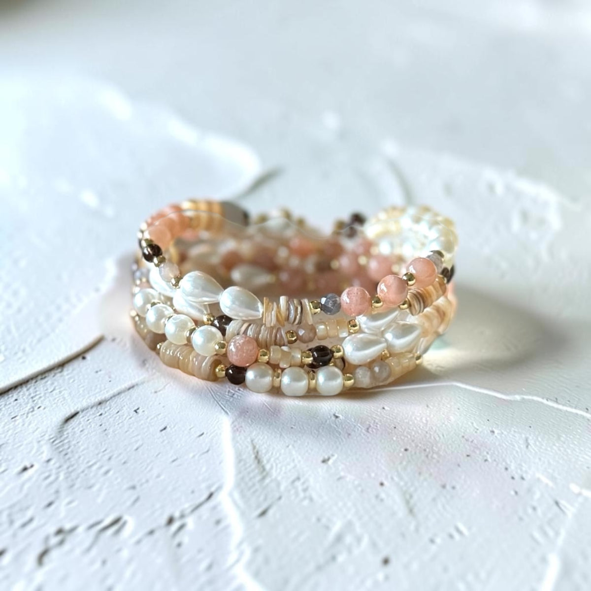 A close-up of a multi-strand beaded bracelet featuring white pearls, peach-pink beads, and metallic gold and silver accents, resting on a textured white surface.