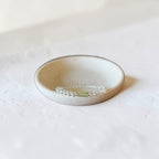 A delicate clear bracelet with a small light green stone accent, displayed inside a small beige ceramic dish on a textured white surface.