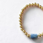 A beaded stretch bracelet featuring metallic gold round beads and a single central light blue rectangular stone, displayed on a textured off-white background.