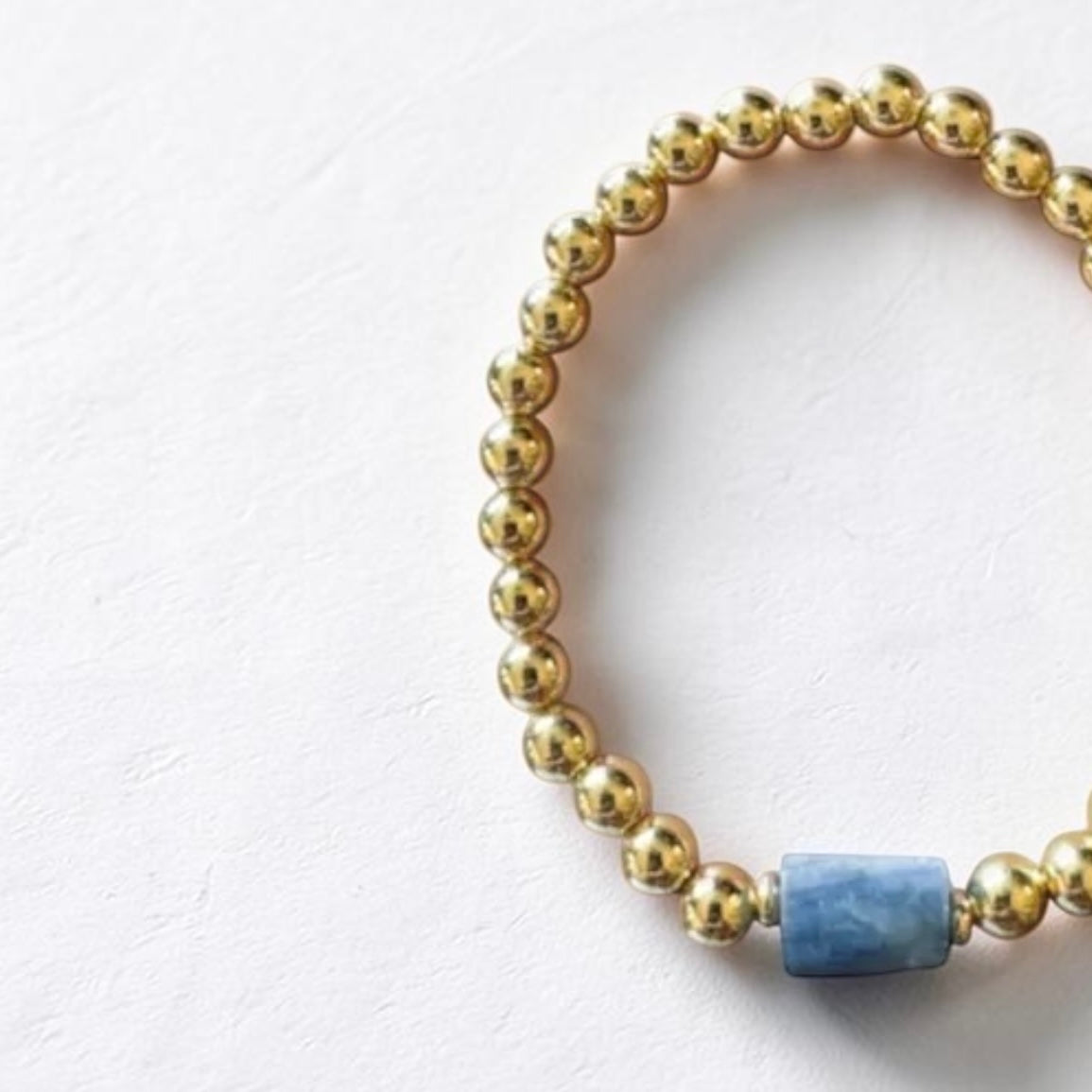 A beaded stretch bracelet featuring metallic gold round beads and a single central light blue rectangular stone, displayed on a textured off-white background.