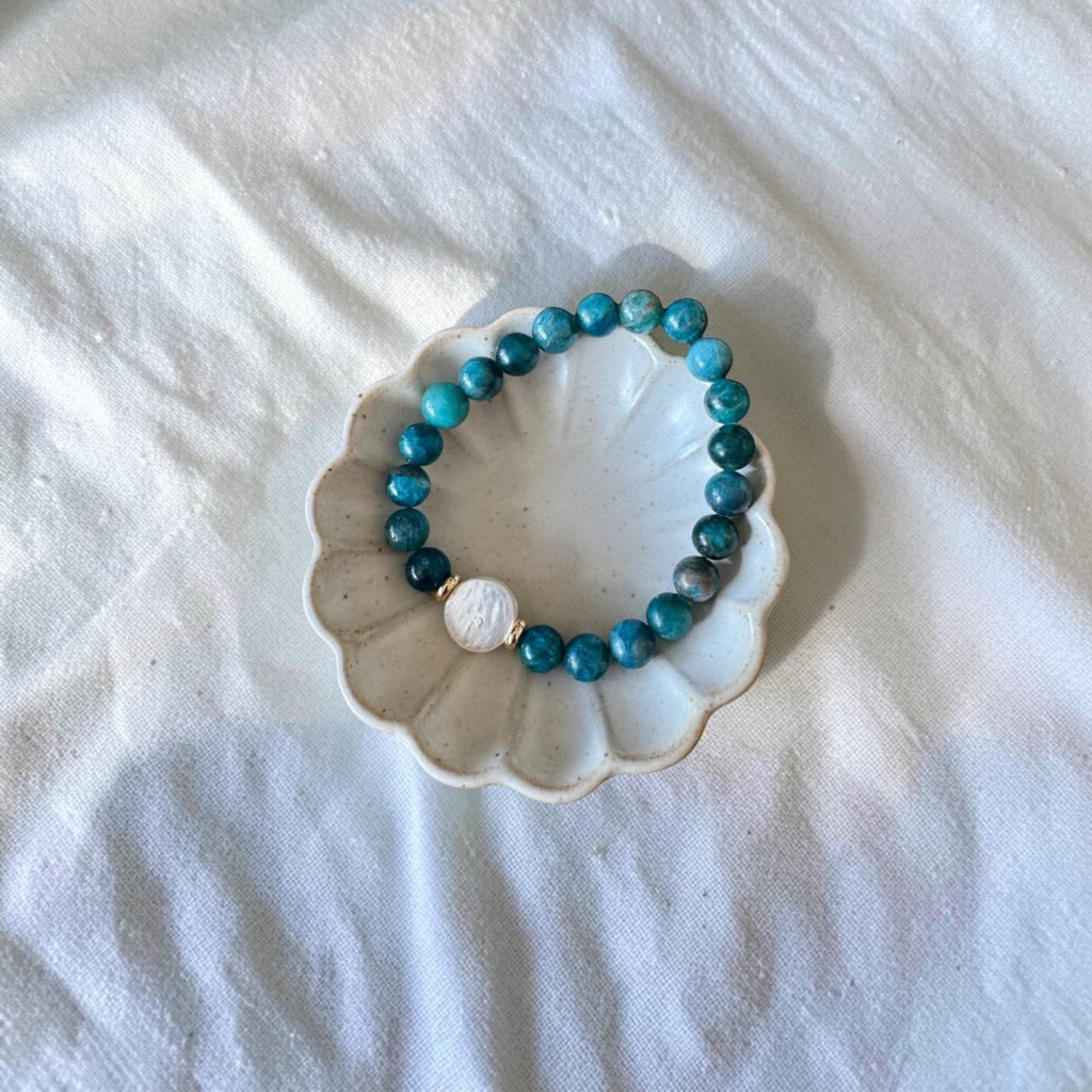 A beaded stretch bracelet with turquoise-blue and dark teal beads, a white round accent bead, and a small gold spacer, displayed on a scalloped white dish over a white fabric background.