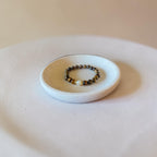 A beaded bracelet with dark brown and black beads and a small gold accent, displayed on a small white ceramic dish.