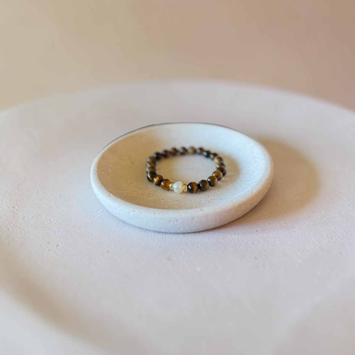 A beaded bracelet with dark brown and black beads and a small gold accent, displayed on a small white ceramic dish.