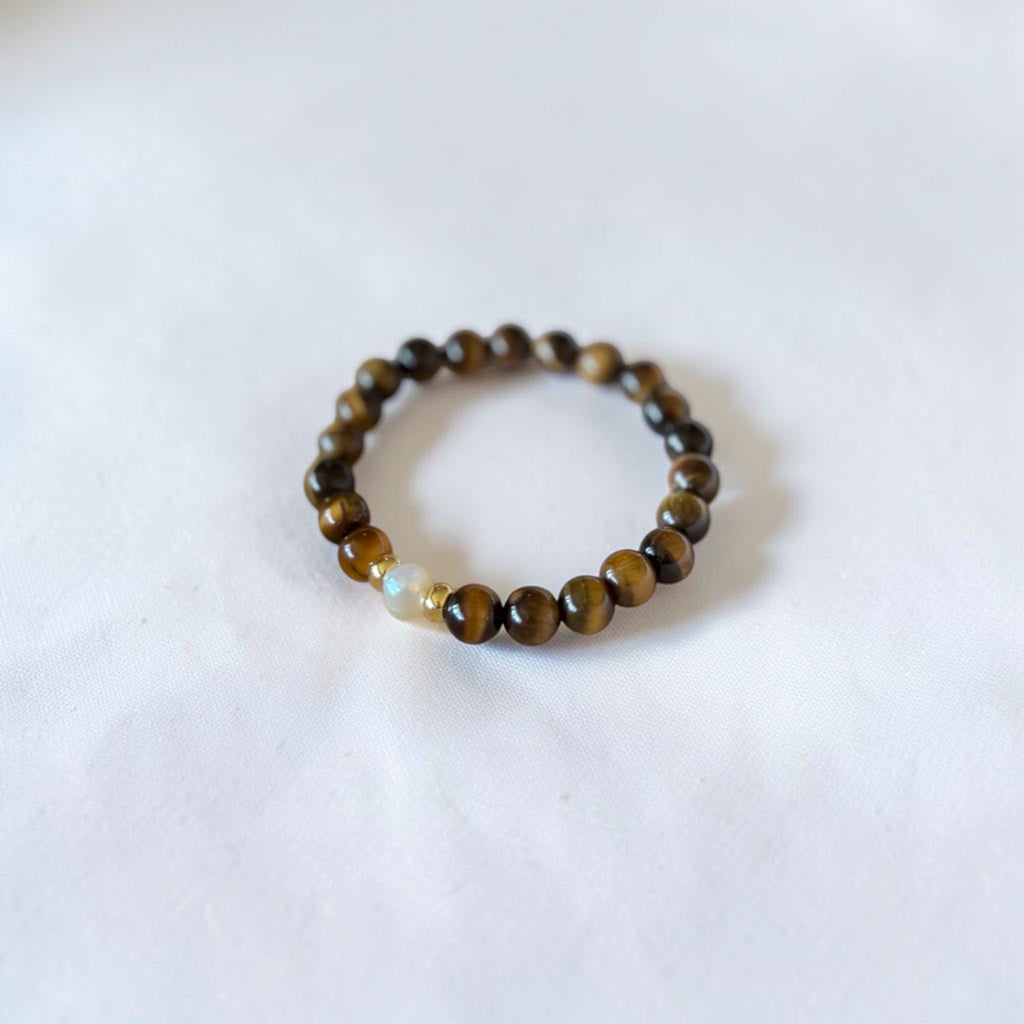 A beaded stretch bracelet featuring round brown and black tiger's eye beads with a single light beige accent bead, displayed on a soft white fabric background.