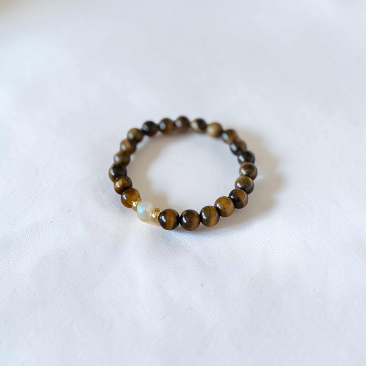 A beaded stretch bracelet featuring round brown and black tiger's eye beads with a single light beige accent bead, displayed on a soft white fabric background.
