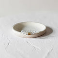 A delicate beaded bracelet with a smalll barrel texture center accent and clear faceted beads, displayed on a small round ceramic dish against a textured light gray background.
