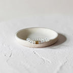 A delicate beaded bracelet with a smalll barrel texture center accent and clear faceted beads, displayed on a small round ceramic dish against a textured light gray background.