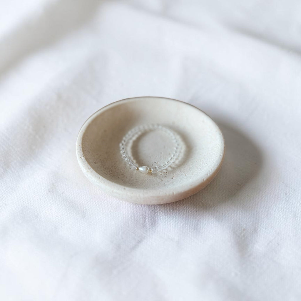 A small round ceramic dish with a textured matte finish, holding a delicate beaded ring, set against a soft white fabric background.