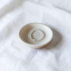 A small round ceramic dish with a textured matte finish, holding a delicate beaded ring, set against a soft white fabric background.